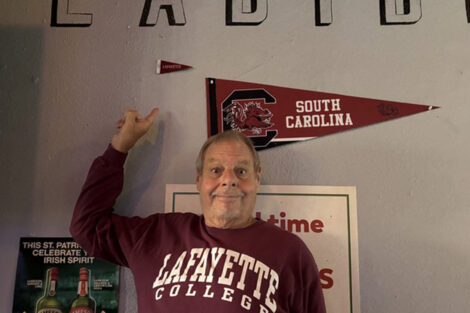An alumnus wearing a Lafayette College sweatshirt points at a flag.