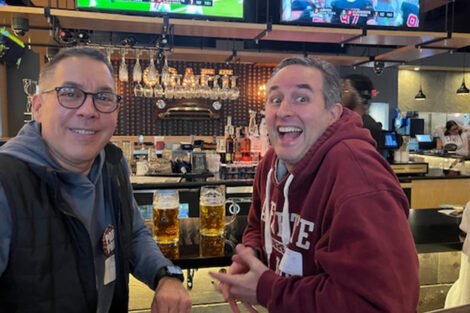 Two men smile at the camera. They are seated at a bar.